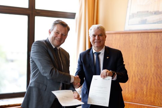 Der Direktor des Kulturzentrums Flagey, Gilles Ledure, und Minister Gregor Freches schütteln sich die Hände und zeigen den unterschriebenen Vertrag