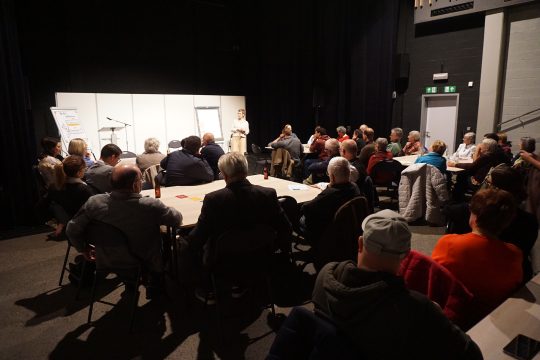 Menschen sitzen in dunklem Saal und hören Vortrag zu