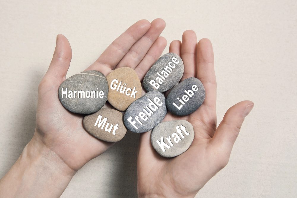 Hand mit Steinen, auf denen die Worte "Harmonie, Mut, Glück, Freude, Balance, Liebe, Kraft" stehen