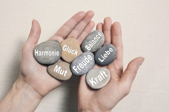 Hand mit Steinen, auf denen die Worte "Harmonie, Mut, Glück, Freude, Balance, Liebe, Kraft" stehen