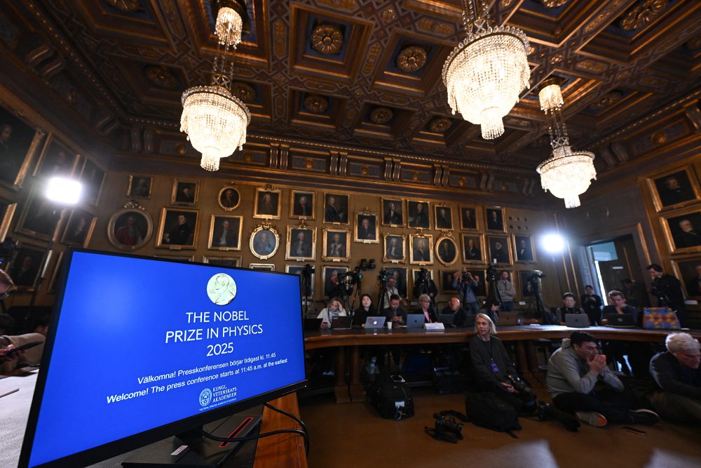 Auf einem Monitor steht in englischer Sprache "Physik-Nobelpreis 2025", in einem Saal warten Journalisten auf den Beginn der Pressekonferenz