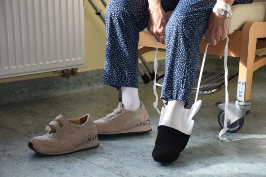 Eine Person sitzt auf einem Stuhl und zieht sich mit einem ergotherapeutischen Hilfsmittel die Schuhe an