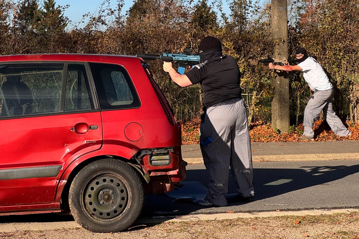 Zwei Männer mit Sturmhauben schießen mit zwei Maschinengewehrattrappen, einer steht hinter einem roten Auto