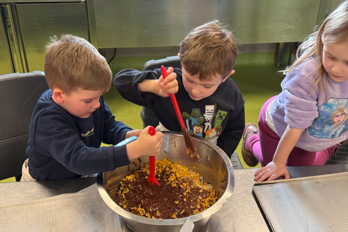 Zwei Jungs rühren Schokoladensauce und Cornflakes in einer Schüssel zusammen, ein Mädchen schaut ihnen zu