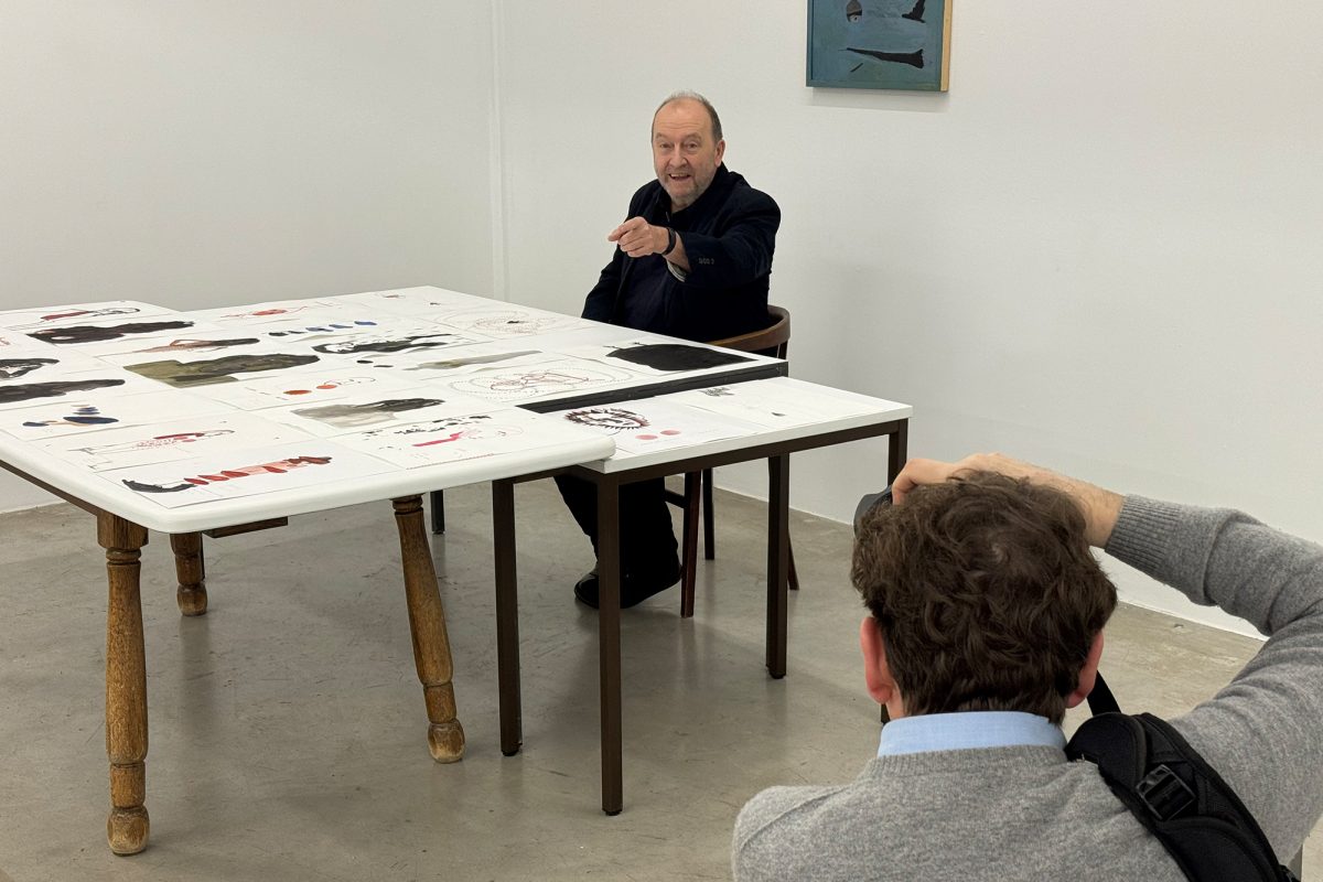 Künstler Ronny Delrue sitzt am Tisch, auf dem mehrere seiner Werke liegen und lacht in die Kamera des Fotografen im Vordergrund