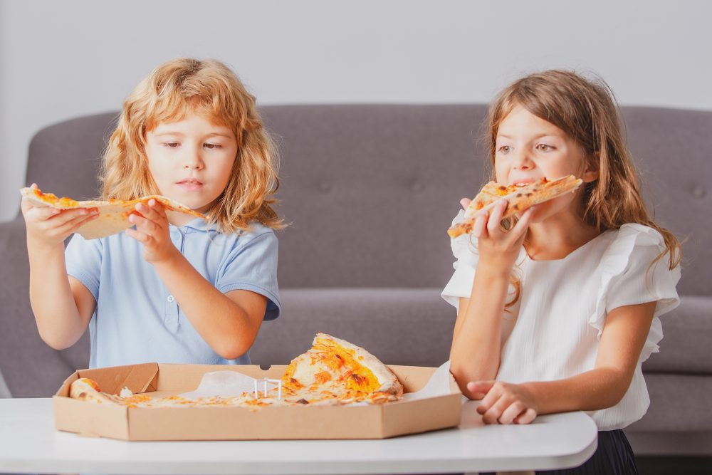 Zwei Mädchen essen Pizza, der Pizzakarton liegt vor ihnen auf einem Couchtisch