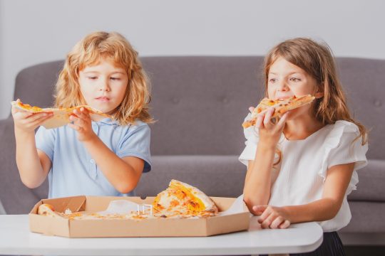 Zwei Mädchen essen Pizza, der Pizzakarton liegt vor ihnen auf einem Couchtisch