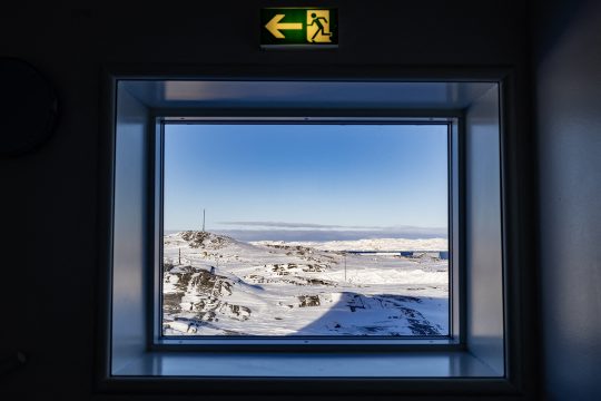 Verschneite Landschaft in Nuuk, Grönland, durch ein Fenster gesehen. Über dem Fenster ein Notausgang-Schild