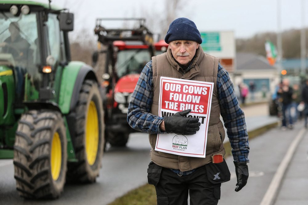 Traktoren auf der Straße, ein Mann trägt ein Plakat mit der Aufschrift "Our cows follow the rules - why don't theirs?" (Unsere Kühe halten sich an die Regeln – warum tun ihre das nicht?)