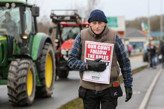 Traktoren auf der Straße, ein Mann trägt ein Plakat mit der Aufschrift "Our cows follow the rules - why don't theirs?" (Unsere Kühe halten sich an die Regeln – warum tun ihre das nicht?)