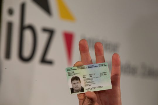 Nahaufnahme einer Hand, die einen belgischen Personalausweis hält, im Hintergrund ist das Logo des Innenministeriums