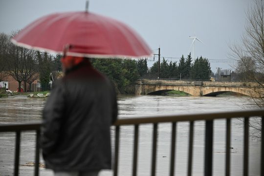 Mann mit Regenschirm steht auf einer Brücke in Coursan, die über den Fluss Aude führt, dessen Pegel stark angestiegen ist