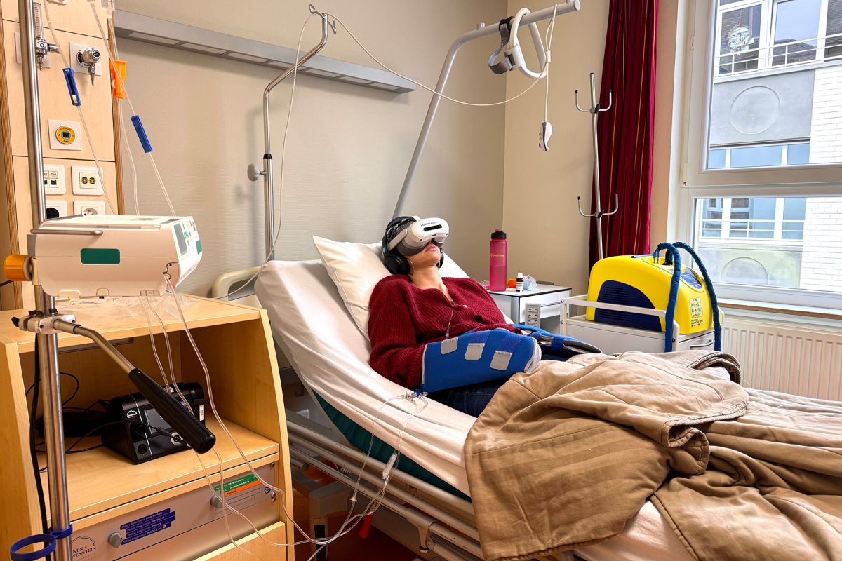 Patientin Morgane Loubris liegt im Krankenhausbett, sie trägt Kühlhandschuhe und die VR-Brille sowie Kopfhörer auf