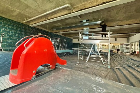 ein Arbeiter auf einem Gerüst arbeitet an der Decke, im Vordergrund eine rote Kinder-Rutschbahn