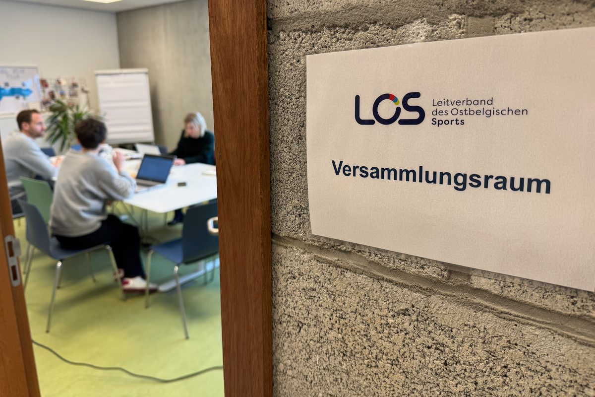 Schild, auf dem "Versammlungsraum" steht, daneben eine geöffnete Tür zu einem Büro, in dem mehrere Personen sitzen