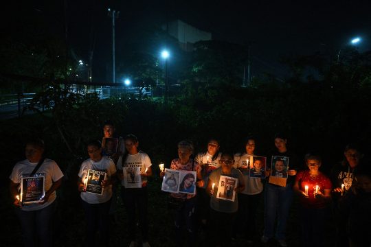 Menschen stehen im Dunkeln vor dem Gefängnis. Sie halten brennende Kerzen und Plakate mit den Bildern inhaftierter Menschen in den Händen