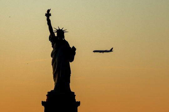 Freiheitsstatue, am orangefarbenen Himmel ist in klein ein Flugzeug im Landeanflug zu sehen