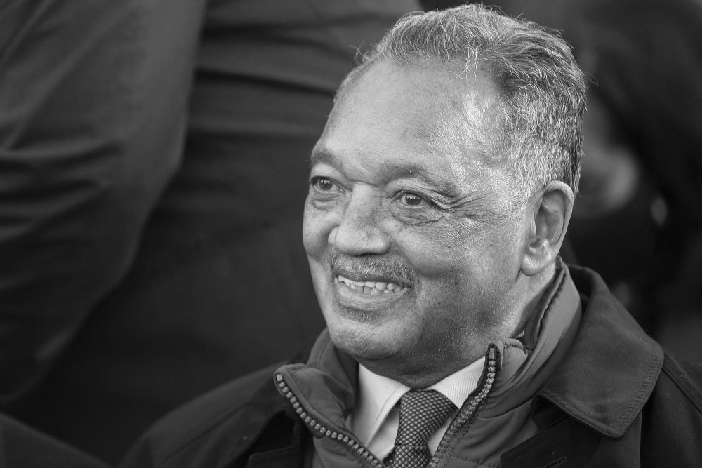 Reverend Jesse Jackson, sitzend, umgeben von stehenden Menschen in Anzügen