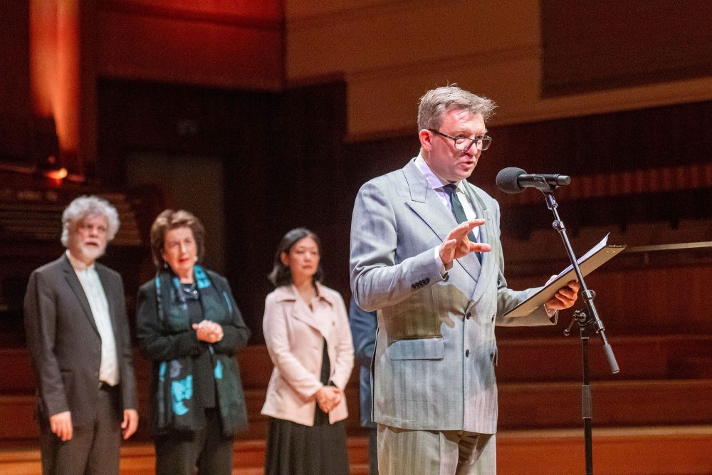 Gilles Ledure am Mikorofon bei der Verkündigung der Resultate des Königin-Elisabeth-Wettbewerbs 2025. Hinter ihm stehen weitere Jury-Mitglieder