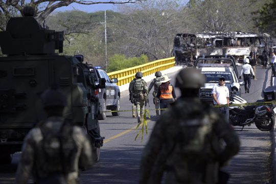 Soldaten in Kampfanzügen und Zivilisten auf der Brücke, im Hintergrund steht ein ausgebrannter Bus quer auf der Fahrbahn