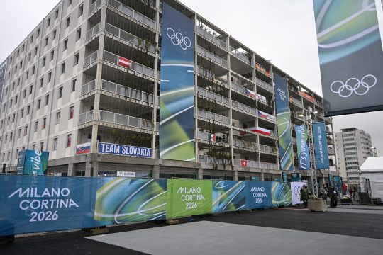 Banner der Winterspiele von Milano-Cortina vor mehrstöckigen Gebäuden des Olympischen Dorfes