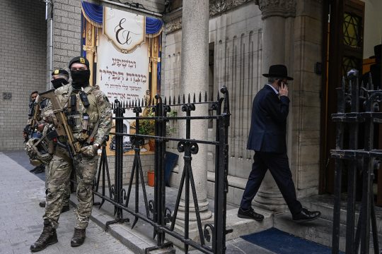 Zwei vermummte Soldaten mit Waffen im Anschlag stehen vor der Synagoge, im Hintergrund telefoniert ein jüdischer Mann mit dem Handy