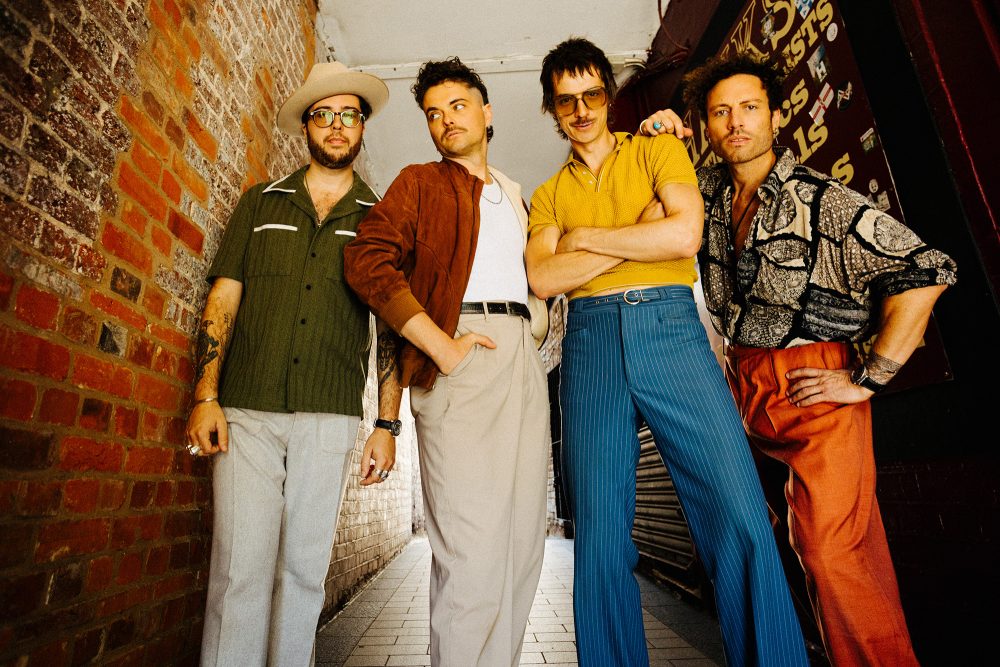 Gruppenbild der vierköpfigen Band in bunter Kleidung vor einer Ziegelwand