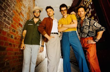 Gruppenbild der vierköpfigen Band in bunter Kleidung vor einer Ziegelwand