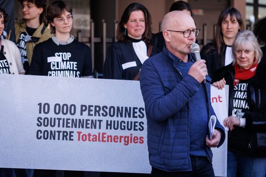 Hugues Falys hält eine Rede neben einem Transparent mit der Aufschrift „10.000 Menschen unterstützen Hugues gegen TotalEnergies“