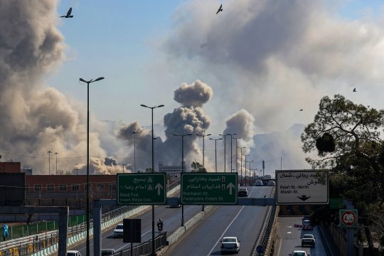 Im Vordergrund eine Autobahn, im Hintergrund Rauchwolken durch Explosionen in Irans Hauptstadt Teheran