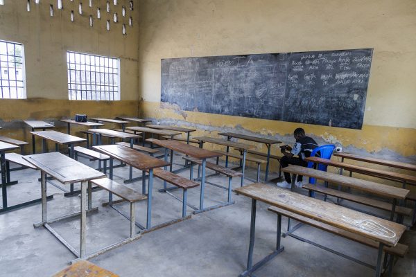 Ein leerer Klassenraum mit Tafel und zahlreichen Pulten. Ein Lehrer sitzt vorn.