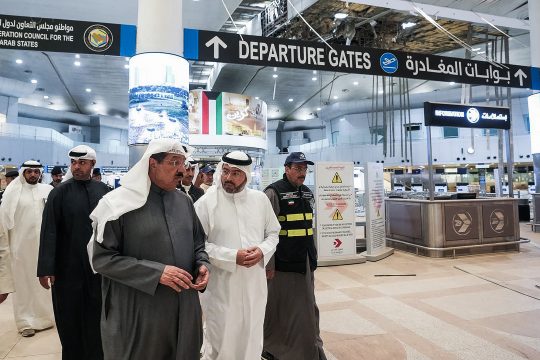 Menschen in der Halle des kuwaitischen Flughafens, im Hintergrund der Zugang zu den Gates, Schilder in englischer und arabischer Sprache