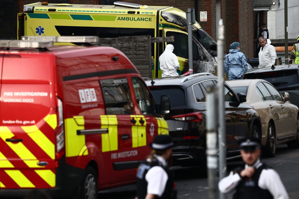 ein Feuerwehrfahrzeug steht am Straßenrand vor einem ausgebrannten Ambulanz-Fahrzeug, mehrerer Forensiker in Schutzanzügen sichern im Hintergrund Spuren, im Vordergrund mehrere Polizisten in Uniform