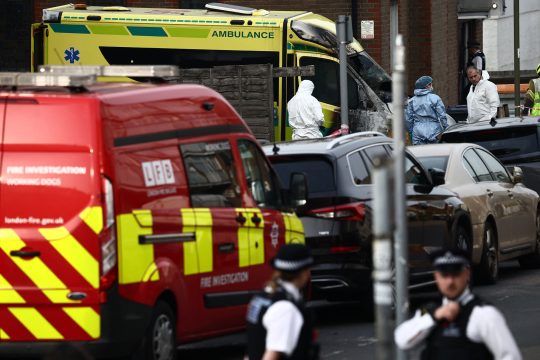 ein Feuerwehrfahrzeug steht am Straßenrand vor einem ausgebrannten Ambulanz-Fahrzeug, mehrerer Forensiker in Schutzanzügen sichern im Hintergrund Spuren, im Vordergrund mehrere Polizisten in Uniform