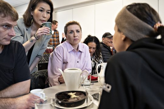 Mette Frederiksen sitzt gemeinsam mit mehreren Frauen und einem Mann an einem Tisch und hört einer Frau zu