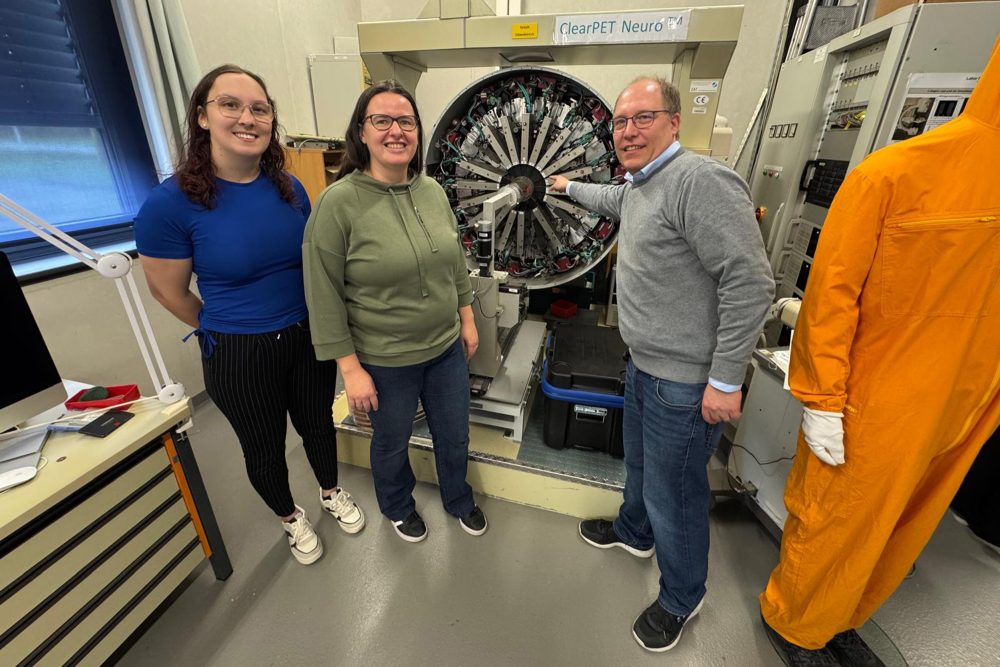 Alina Kaulen, Elisabeth Paulßen und Christoph Langer stehen am Campus Jülich neben einer lebensgroßen orangefarbenen Puppe und vor einer Maschine zur bildgebenden Diagnostik