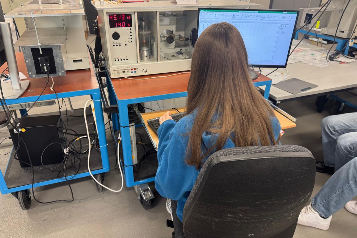 Studentin am Campus Jülich der FH Aachen, von hinten, sie sitzt auf einem Bürostuhl an einem Arbeitstisch, auf dem ein PC und eine wissenschaftliche Apparatur zu finden sind