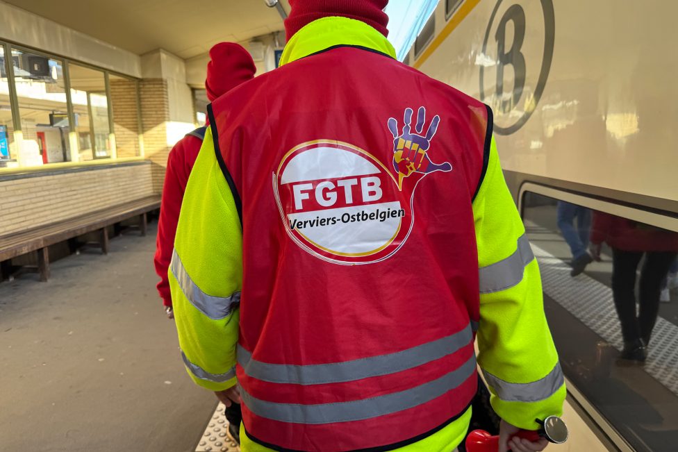 Person von hinten auf einem Bahnhof mit roter Weste mit Aufschrift "FGTB Verviers-Ostbelgien"
