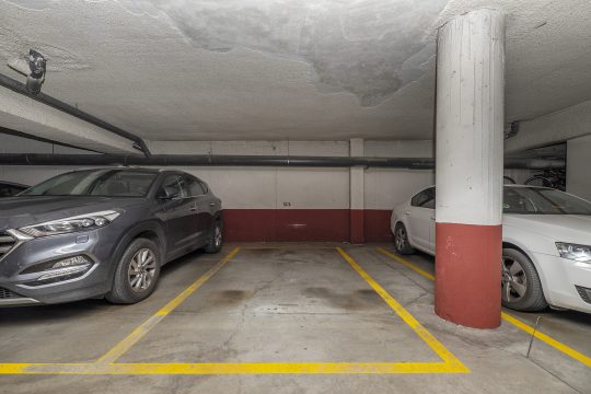 Ein in die Jahre gekommenes Parkhaus mit niedriger Decke, an der der Putz abgebröckelt ist. Zwei Autos sind geparkt. Zwischen ihnen ist eine leere Parklücke.