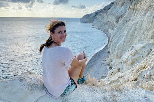 Syna Gensterblum sitzt an den Klippen der Küste von Zypern und lächelt in die Kamera