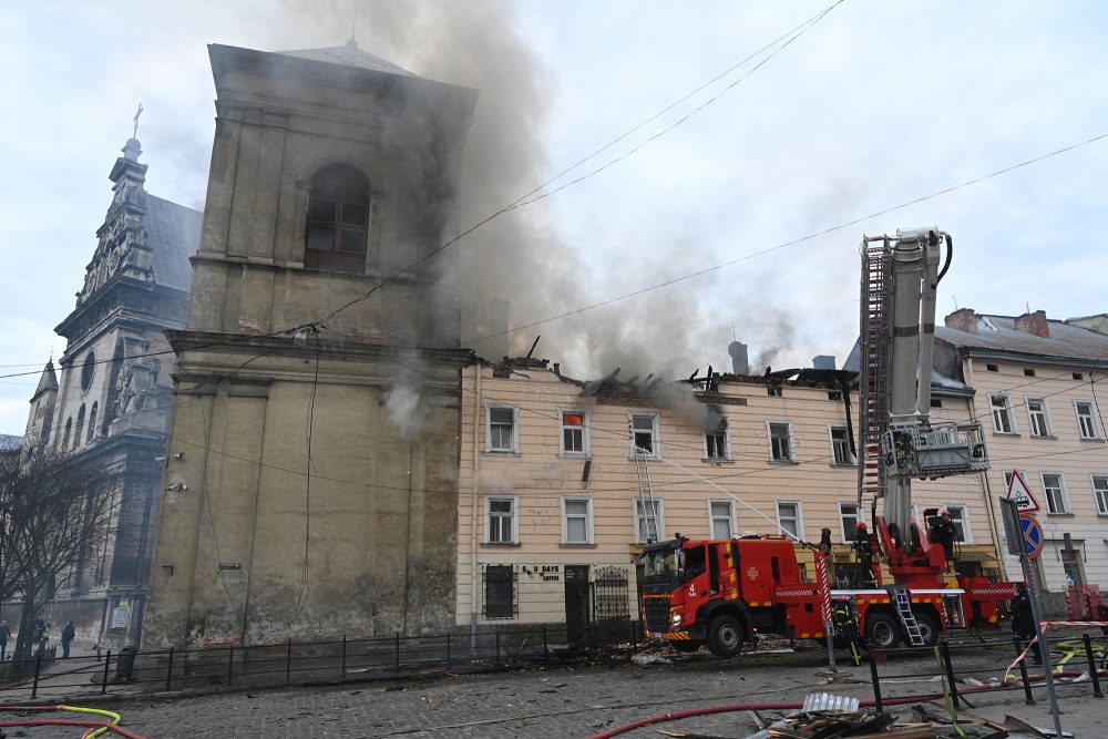 Rauch steigt aus einem mehrstöckigen Gebäude in der Nähe einer alten Kirche in Lwiw auf, Feuerwehrleute sind mit Löschen beschäftigt. Vor dem Gebäude steht ein Feuerwehrfahrzeug mit Hebebühne.