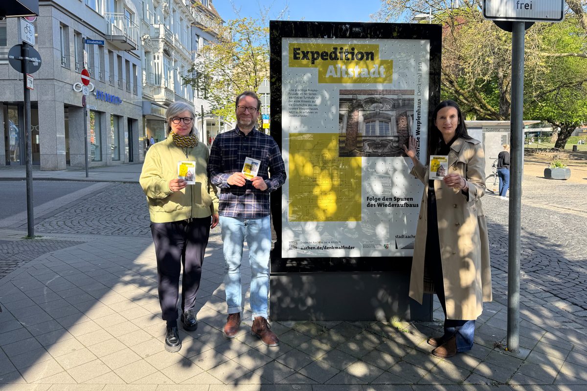 Stadtbaurätin Frauke Burgdorff, Andreas Priesters von der Abteilung Denkmalpflege und Archäologie und Stadtkonservatorin Monika Krücken stehen vor einem Plakat-Citylight, auf dem Werbung für die "Expedition Altstadt" gemacht wird, außerdem halten sie entsprechende Flyer in den Händen