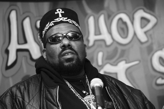 Afrika Bambaataa mit Mikro in der Hand bei einer Pressekonferenz, im Hintergrund der Schriftzug "Hip-Hop"
