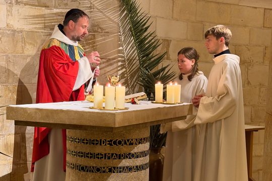 Abt Nikodemus Schnabel bei der Feier am Palmsonntag in der Abtei Dormitio in Jerusalem, er steht hinter dem Altar und hat die Hände zum Gebet gefaltet, zwei Messdiener stehen neben ihm