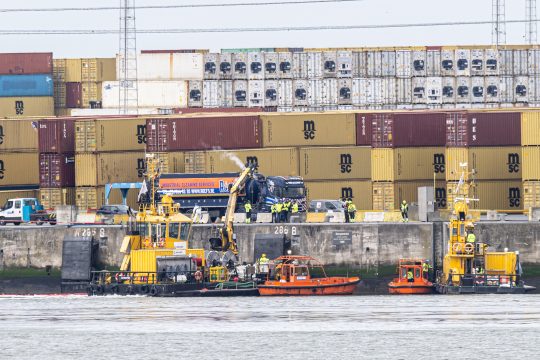 Reinigungsarbeiten werden durchgeführt, ein spezielles Schiff pumpt Öl in einen Laster, im Hintergrund stehen Container gestapelt aufeinander