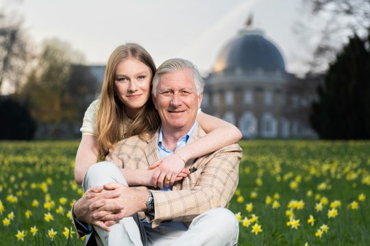 König Philippe und Prinzessin Eléonore sitzen auf einer Blumenwiese vor dem Palast und lächeln in die Kamera