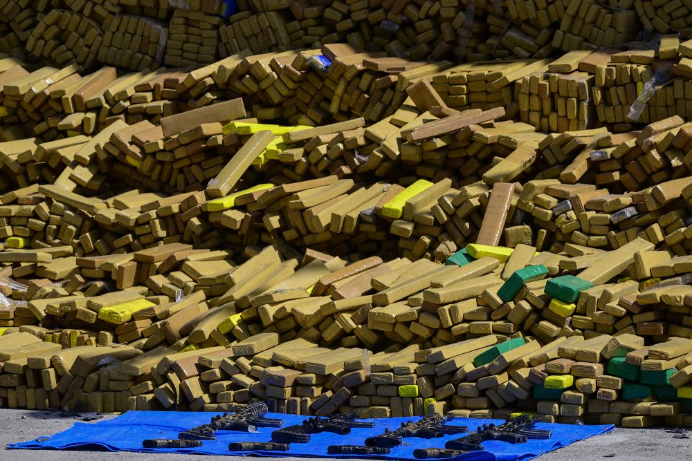 Eine Vielzahl von Marihuana-Päckchen liegen gestapelt aufeinander, davor liegen mehrere beschlagnahmte Maschinenpistolen auf dem Boden