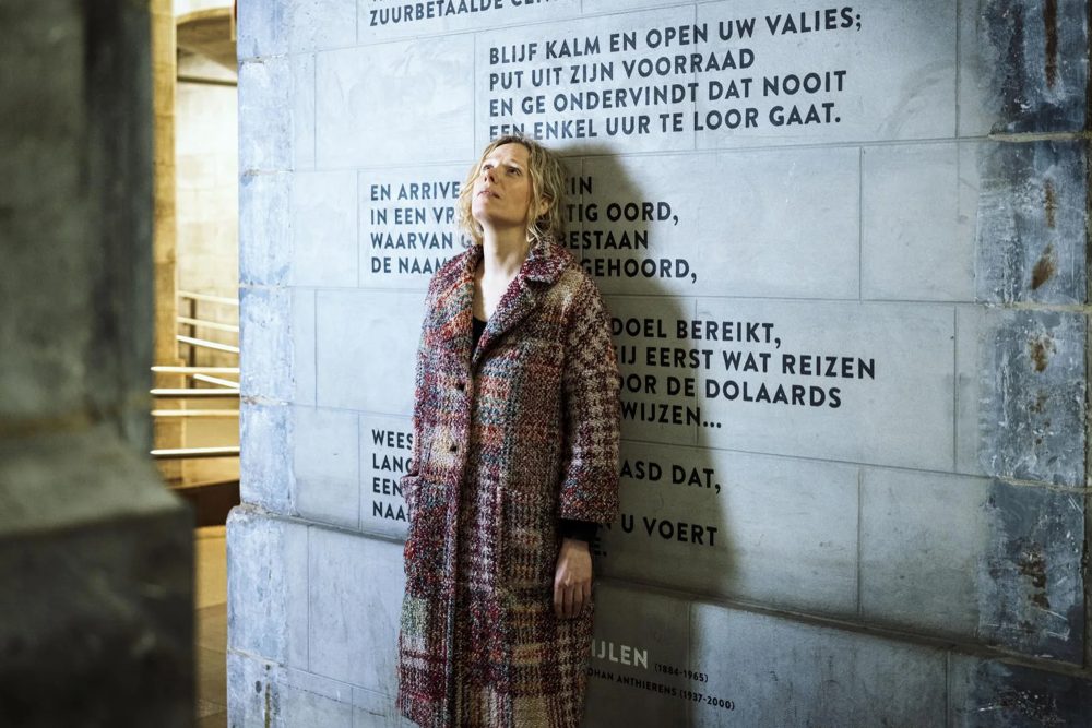 Ruth Lasters steht vor einer Mauer, auf der ein flämischsprachiger Text steht