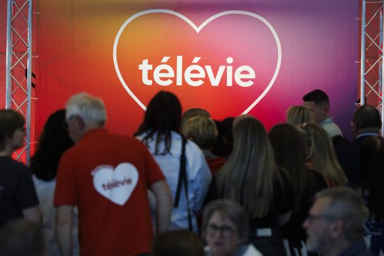 Logo der Spendenaktion Télévie an einer Wand, davor stehen Menschen