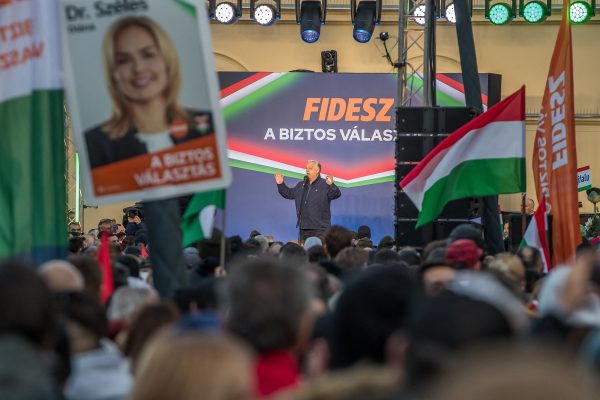 Viktor Orban spricht von einer Bühne aus zu einer großen Menschenmenge. Im Hintergrund das Logo und der Schriftzug der Fidesz-Partei.